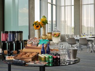 An Assortment Of Snacks, Pastries, Coffee, Beverages, Cups And Ice On A Table With A Meeting Room Setup In The Background